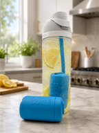 A clear insulated water bottle with a white lid stands on a kitchen counter, filled with lemon slices and carbonated liquid. A Frosty Core Cooling Core and blue straw are inside the water bottle and another Frosty Core is sitting on the counter.